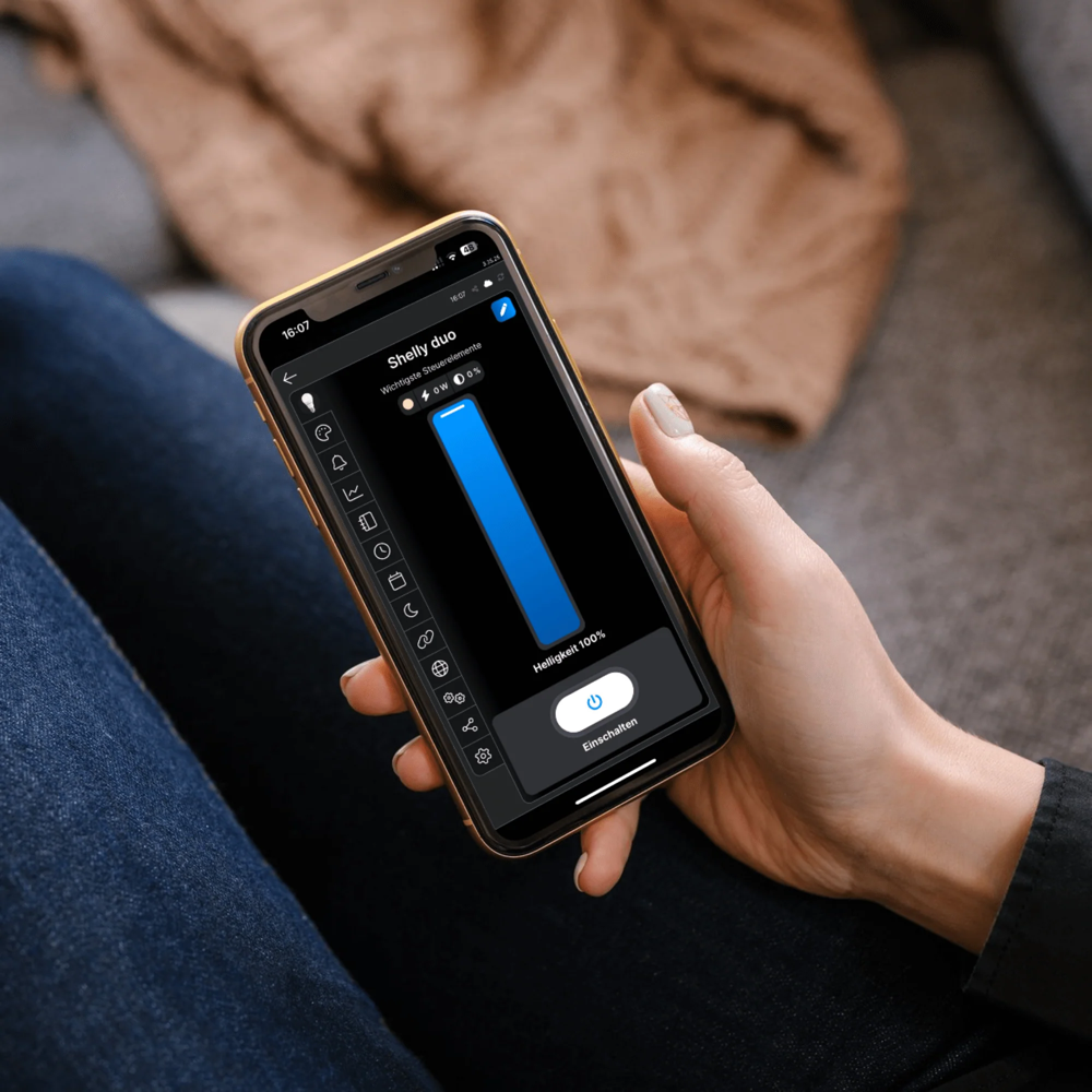 A person holding a smartphone showing the Shelly Cloud app interface used to dim a Shelly smart bulb at 100% brightness.